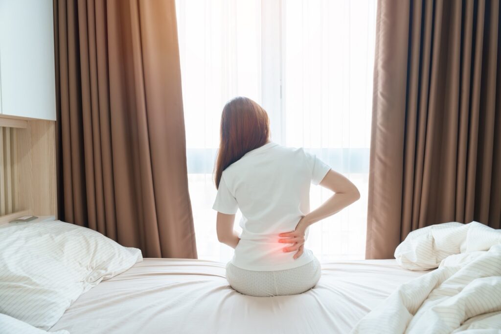Woman sitting on the edge of a bed holding her lower back, with a red highlight indicating low back pain
