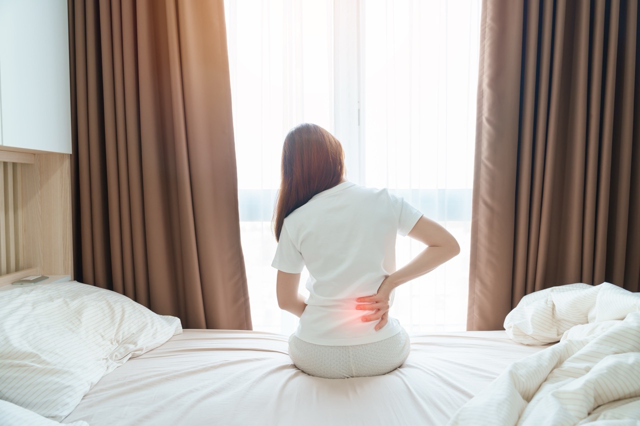 Woman sitting on the edge of a bed holding her lower back, with a red highlight indicating low back pain